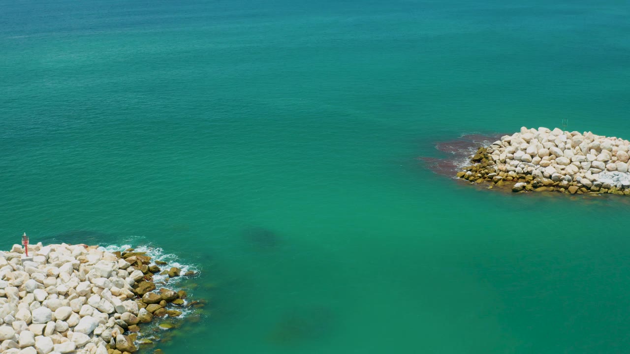 Aerial frame from San Jose Del Cabo, Mexico, with a cropped rock breakwater at the lower left, another pale stone pile at upper right, and open turquoise water in between, with no visible people.