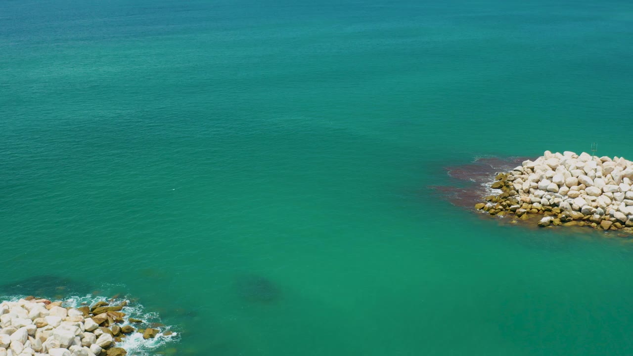 A pale rock mound sits near the upper right of green-blue ocean water in San Jose Del Cabo, Mexico, with a smaller rock edge entering from below and no visible people.