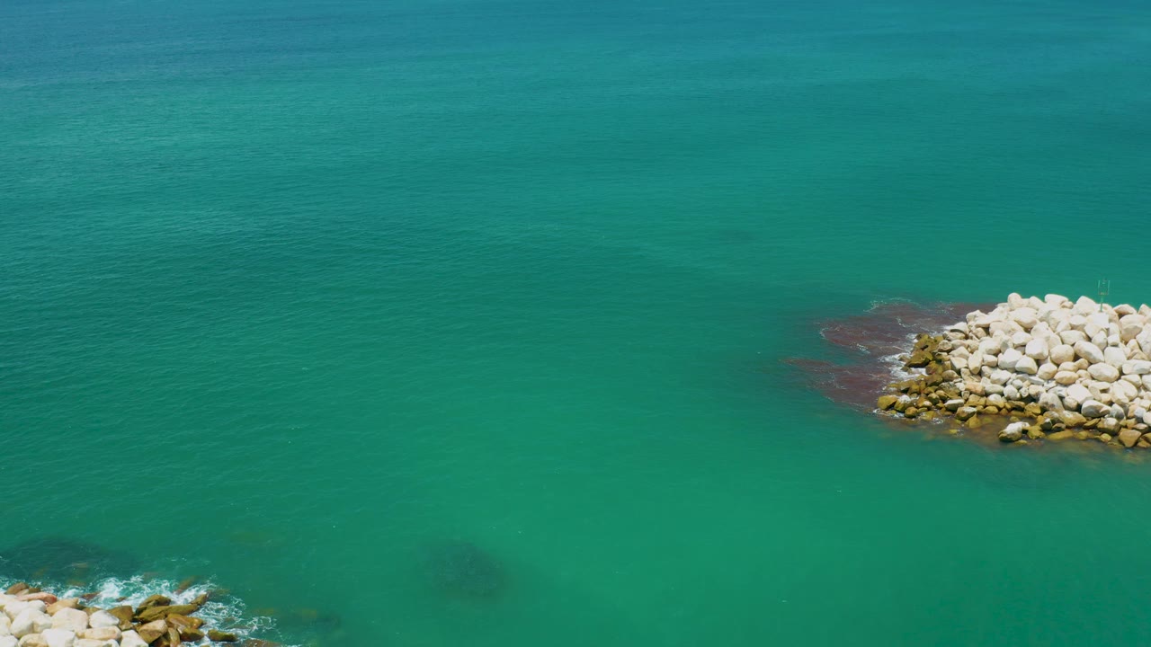 Green-blue ocean fills the frame near San Jose Del Cabo, Mexico, with a pale rock mound on the right side, a smaller rock edge below, and no visible people.