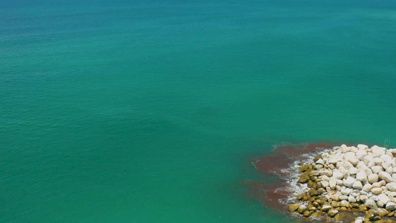 Clear turquoise water surrounds a pale rocky corner in San Jose Del Cabo, with reddish shallows visible around the stones and no visible people.