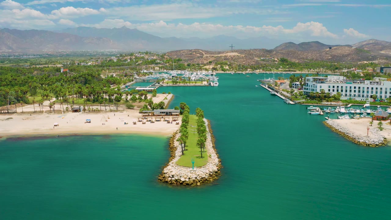 Opening aerial frame over San Jose Del Cabo with turquoise water in front, a narrow palm-lined breakwater centered, sandy beach and marina behind it, distant hills and mountains, and no visible people.