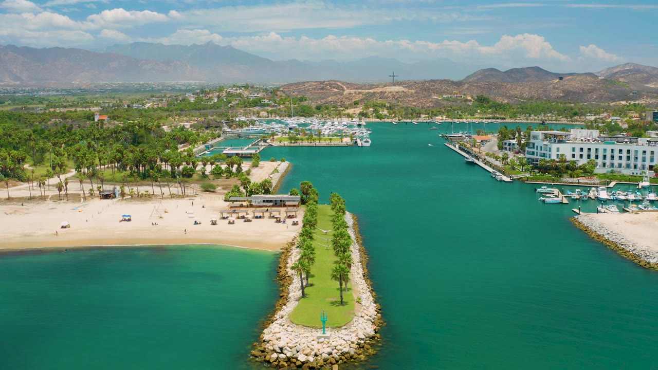San Jose Del Cabo aerial view with the narrow palm breakwater centered low in frame, beach structures to the left, marina boats to the right, hills beyond town, and no visible people.