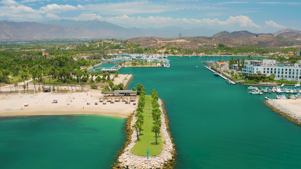 Wide San Jose Del Cabo aerial frame showing turquoise marina water, a sandy beach with small distant objects, a palm-lined point, white waterfront buildings, mountains, and no visible people.