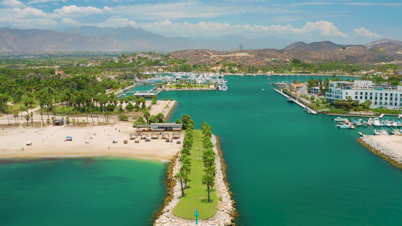 San Jose Del Cabo frame looking over green-blue coastal water toward the sandy beach, marina slips, white buildings on the right, palm rows, low hills, and no visible people.