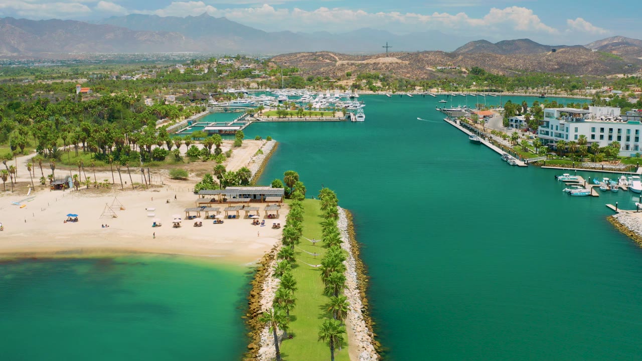 Aerial frame over San Jose Del Cabo showing turquoise water around a sandy beach, a palm-lined breakwater in the foreground, docks to the right, white resort buildings, hills, and no visible people.