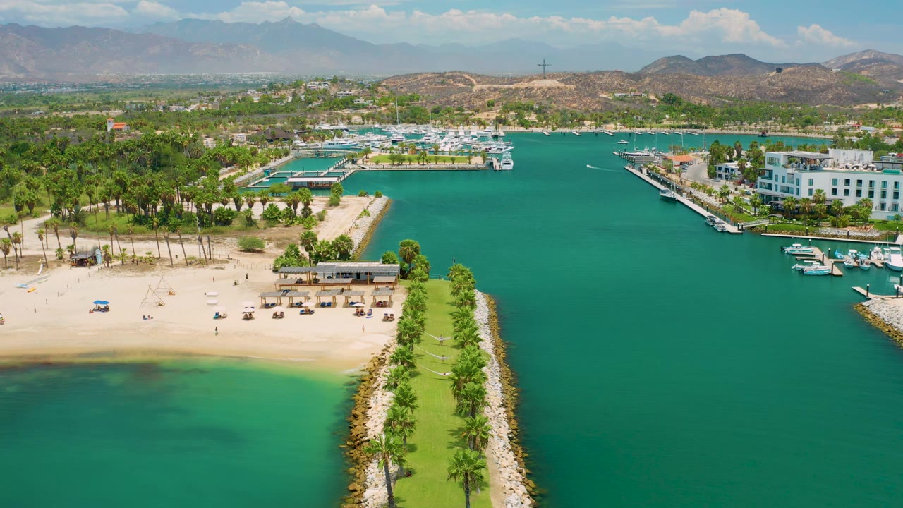 Aerial view in San Jose Del Cabo with the palm breakwater centered lower in frame, a sandy beach to the left, deep turquoise marina water to the right, resort buildings, hills, and no visible people.