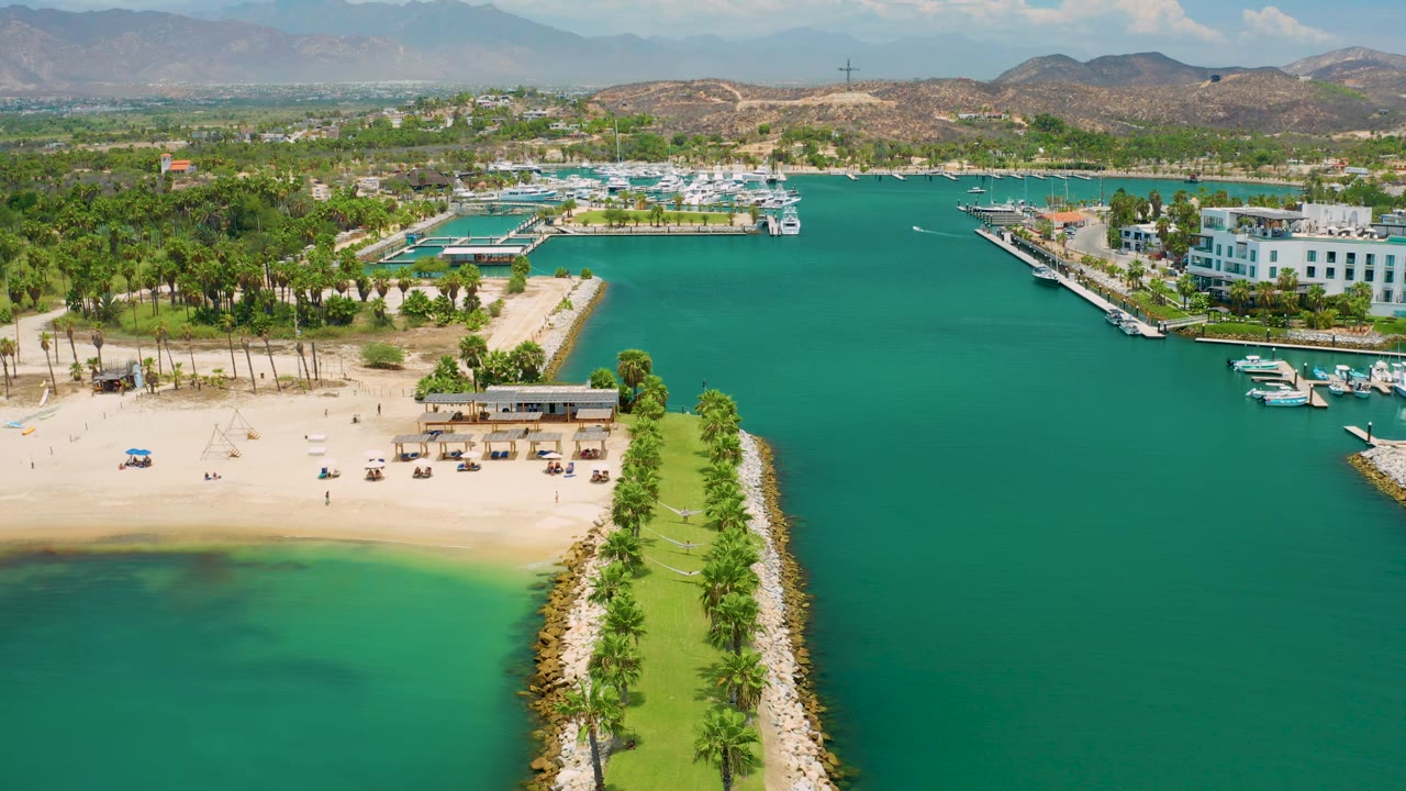 San Jose Del Cabo aerial frame with more open turquoise water on the right, the sandy beach and palm grove on the left, white resort buildings near docks, distant hills, and no visible people.