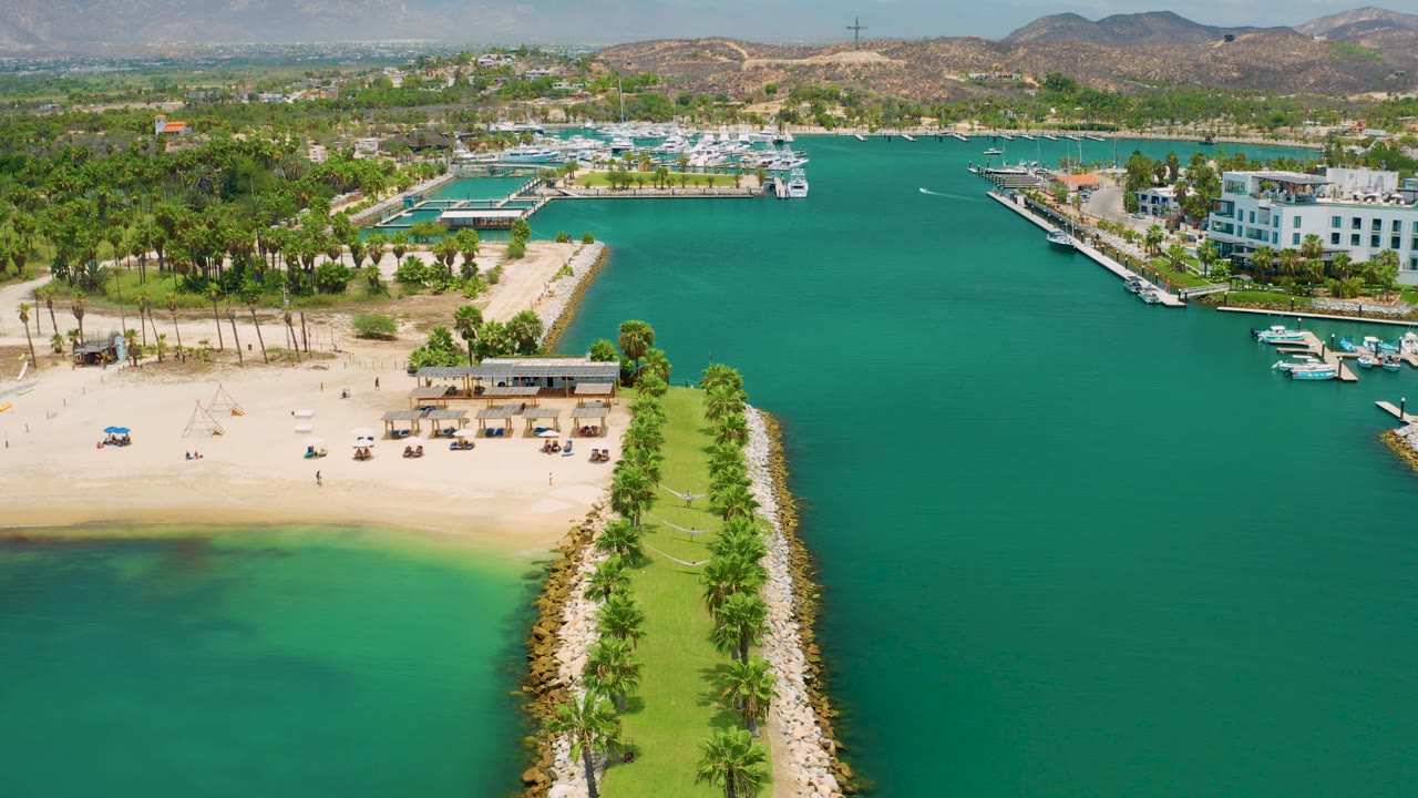 Aerial view over San Jose Del Cabo showing the sandy beach across the upper left, the palm breakwater dropping through the lower right, marina docks, resort buildings, hills, and no visible people.