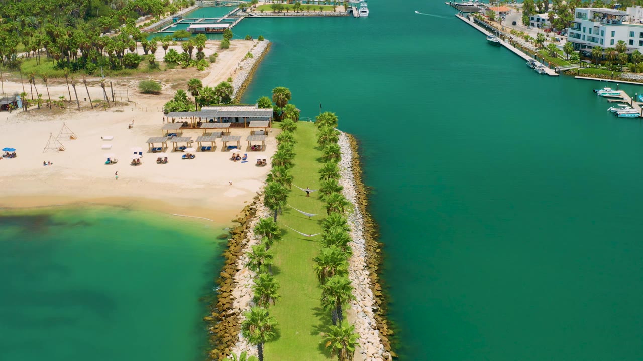 Aerial view in San Jose Del Cabo, Mexico with the jetty centered, hammocks visible along the grass, beach shade structures near the sand, boats by docks, and no visible people.