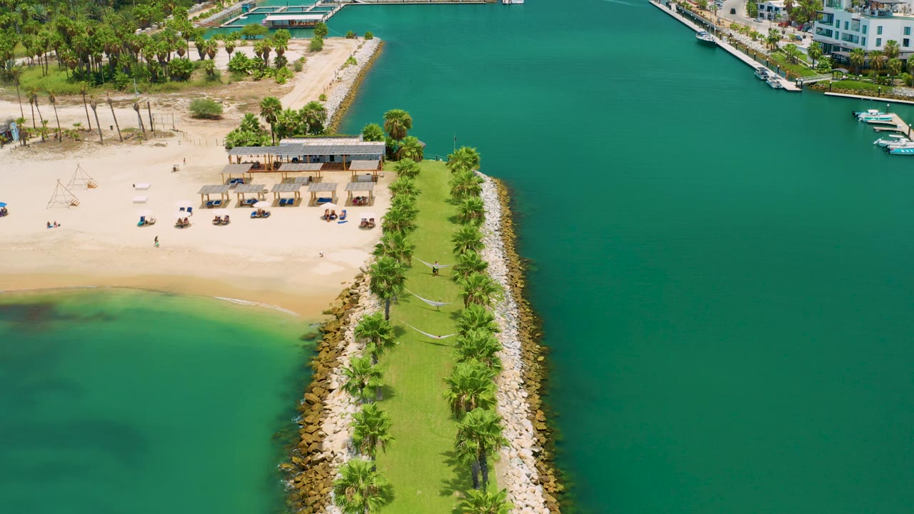 High aerial frame over San Jose Del Cabo, Mexico showing a narrow landscaped jetty bordered by rocks, calm green water on the right, shallow shoreline water on the left, and no visible people.