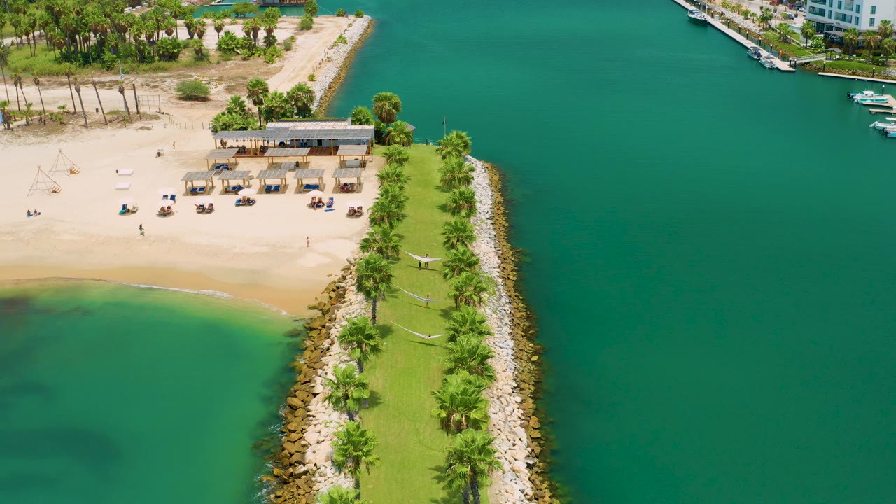 Aerial frame from San Jose Del Cabo, Mexico showing beach cabanas or shade frames, a low beach building, the palm-lined jetty curving through the foreground, turquoise water, and no visible people.