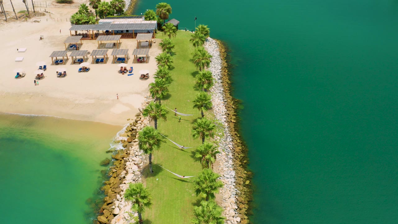 Aerial frame from San Jose del Cabo showing shade huts and lounge chairs on pale sand, a palm-lined grassy strip, rock edging, and green-blue water on both sides with tiny people visible.