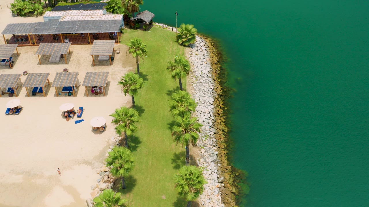 Overhead view in San Jose Del Cabo, Mexico, showing sandy resort seating, green lawn, palm trees, a rock shoreline, and green-blue water with no visible people.