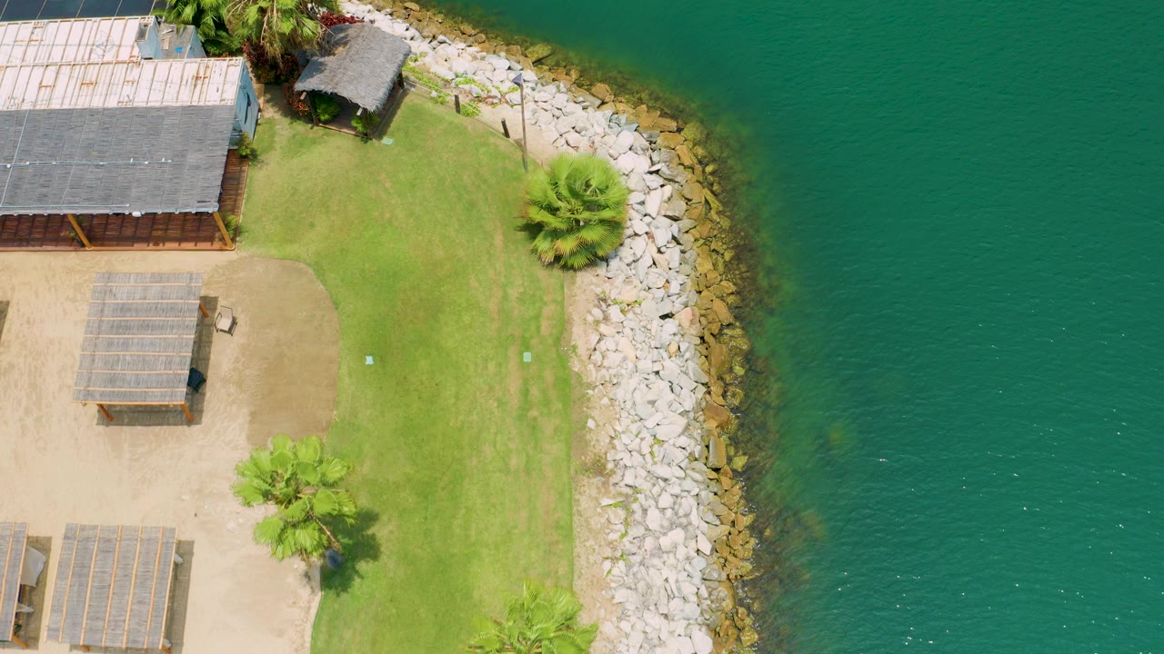 Overhead aerial frame in San Jose del Cabo, Mexico, showing a grassy waterfront campground, shaded structures, a curved rock edge, turquoise water, and no visible people.