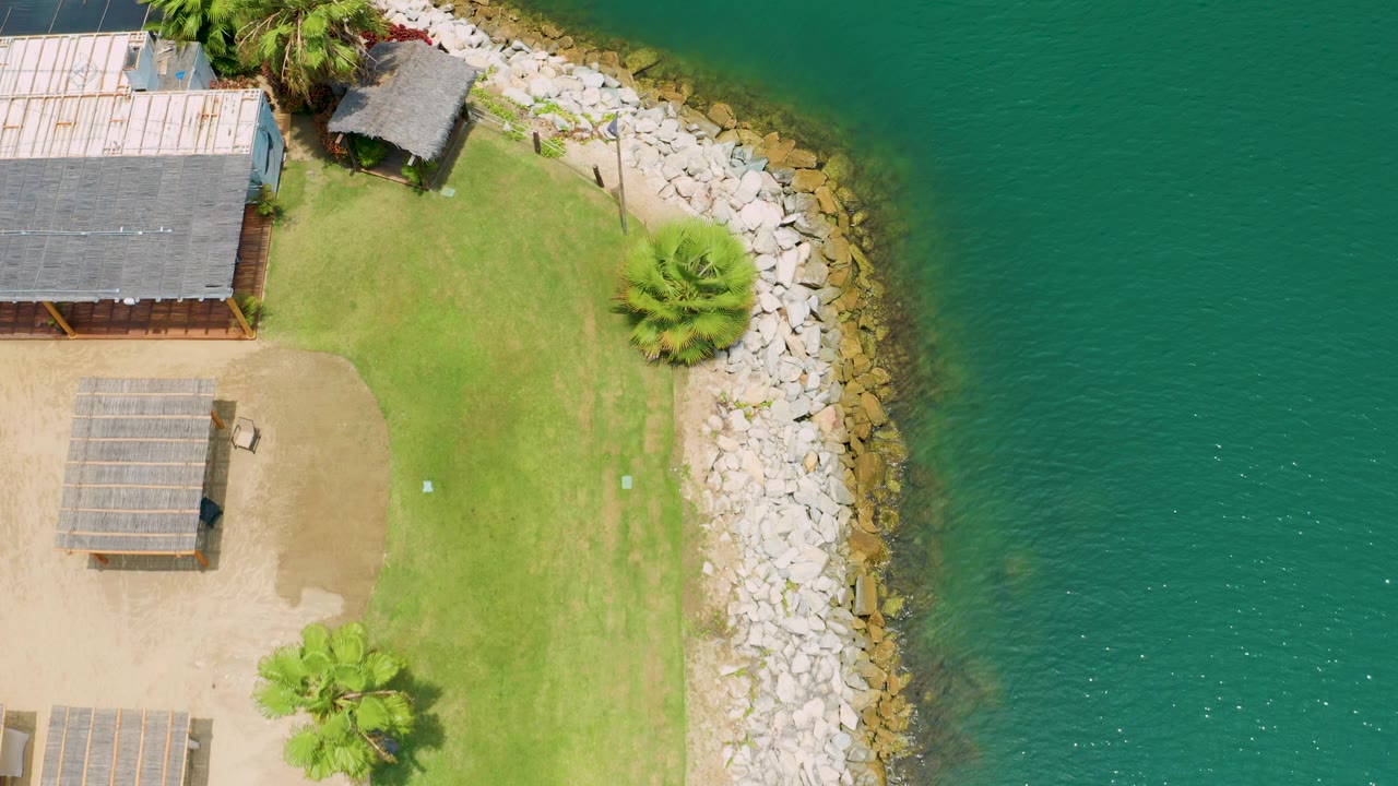 Top-down frame in San Jose del Cabo, Mexico, showing more open turquoise water to the right, a curved rock barrier, trimmed grass, palm trees, and no visible people.