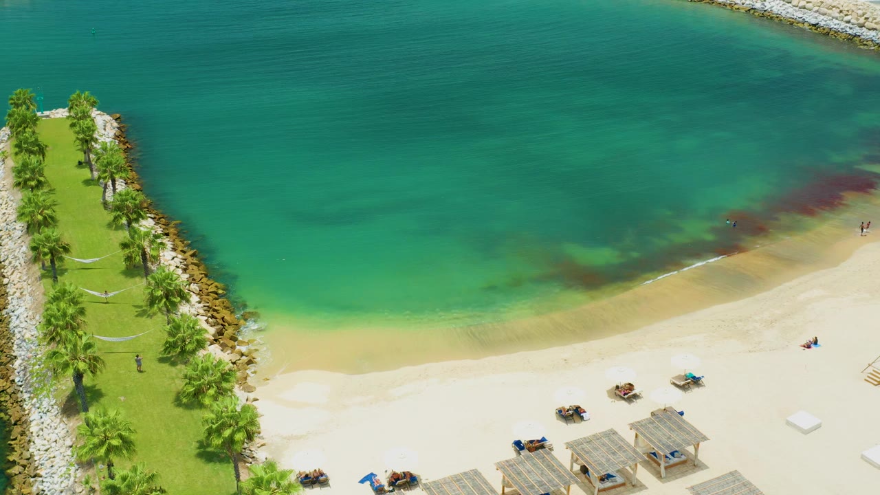 Overhead frame from San Jose Del Cabo showing turquoise sea, pale beach, a palm-lined park, a rock breakwater, lounge chairs, and tiny beachgoers near the shore.
