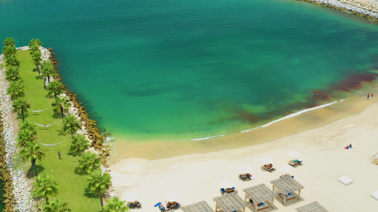 The aerial view over San Jose Del Cabo shows light surf along the sand, dark patches under the water, a palm park at left, and small beachgoers spread along the beach.