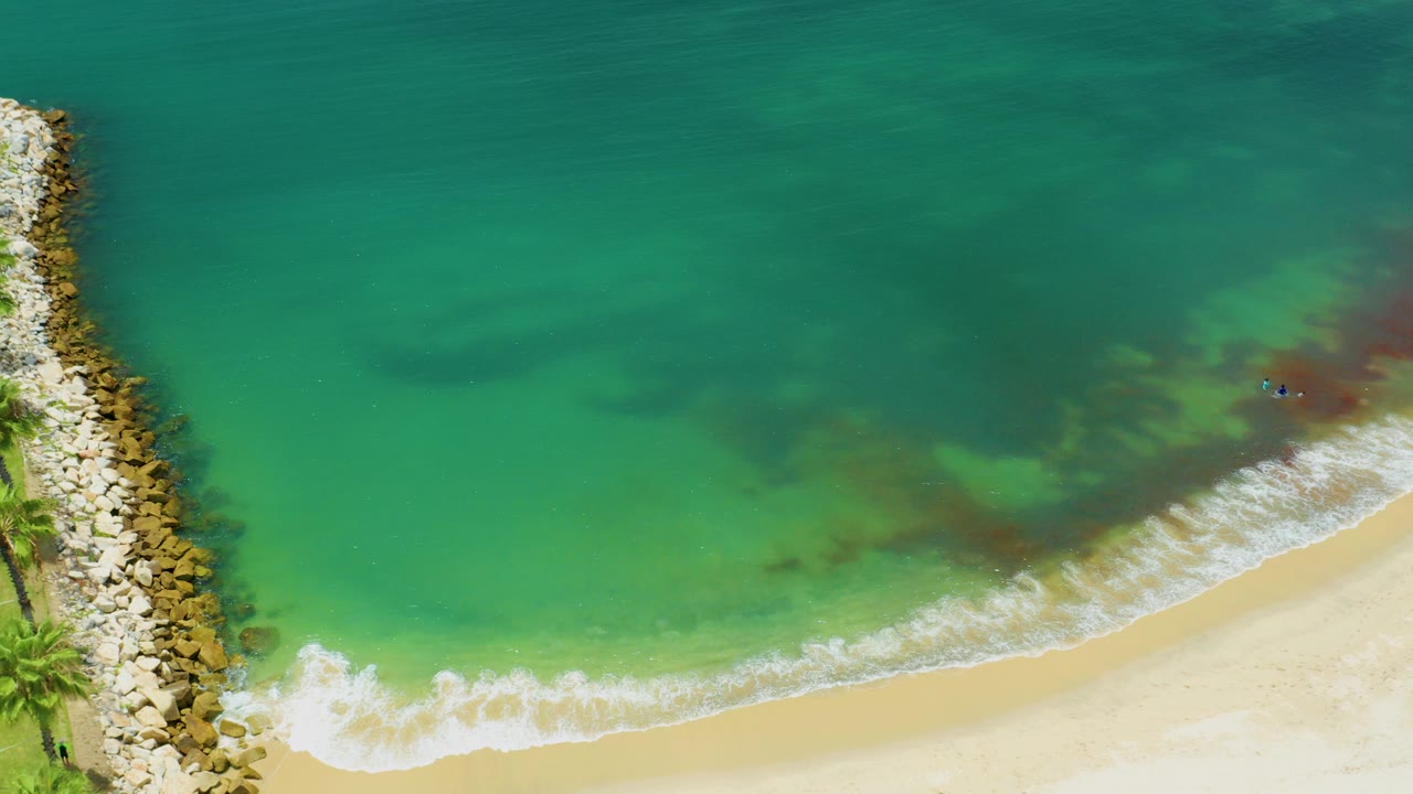Overhead drone frame of San Jose del Cabo shoreline with turquoise water, pale sand, white surf, a rock breakwater on the left, and no visible people.