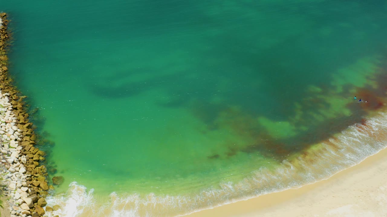Drone view of the San Jose del Cabo coast with the rock breakwater near the middle, surf folding onto sand, turquoise water, and no visible people.
