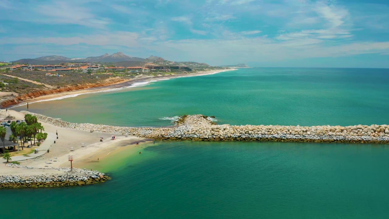 Wide aerial frame from San Jose del Cabo, Mexico, showing teal water, a pale sandy beach, a long surf line, rock breakwaters in the foreground, distant hills, and no visible people.