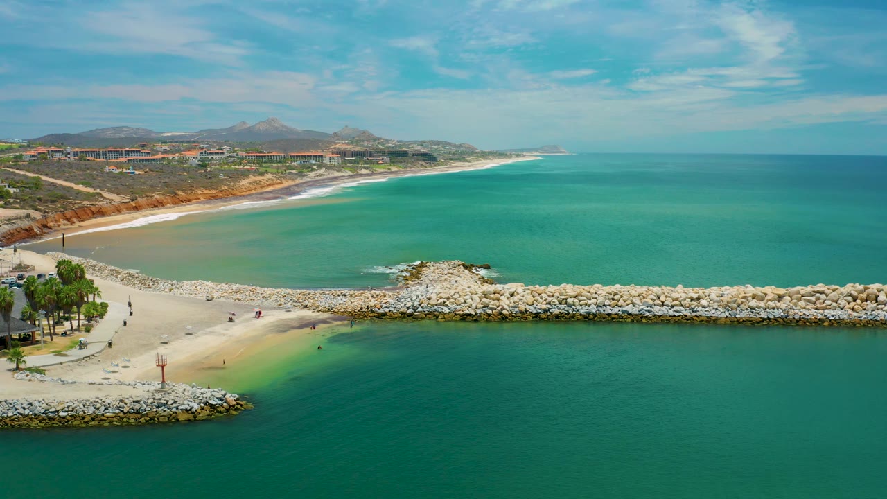 Drone view over San Jose del Cabo, Mexico, with green-blue water filling the right side, a curved beach and rocky breakwater below, inland hills on the horizon, and no visible people.