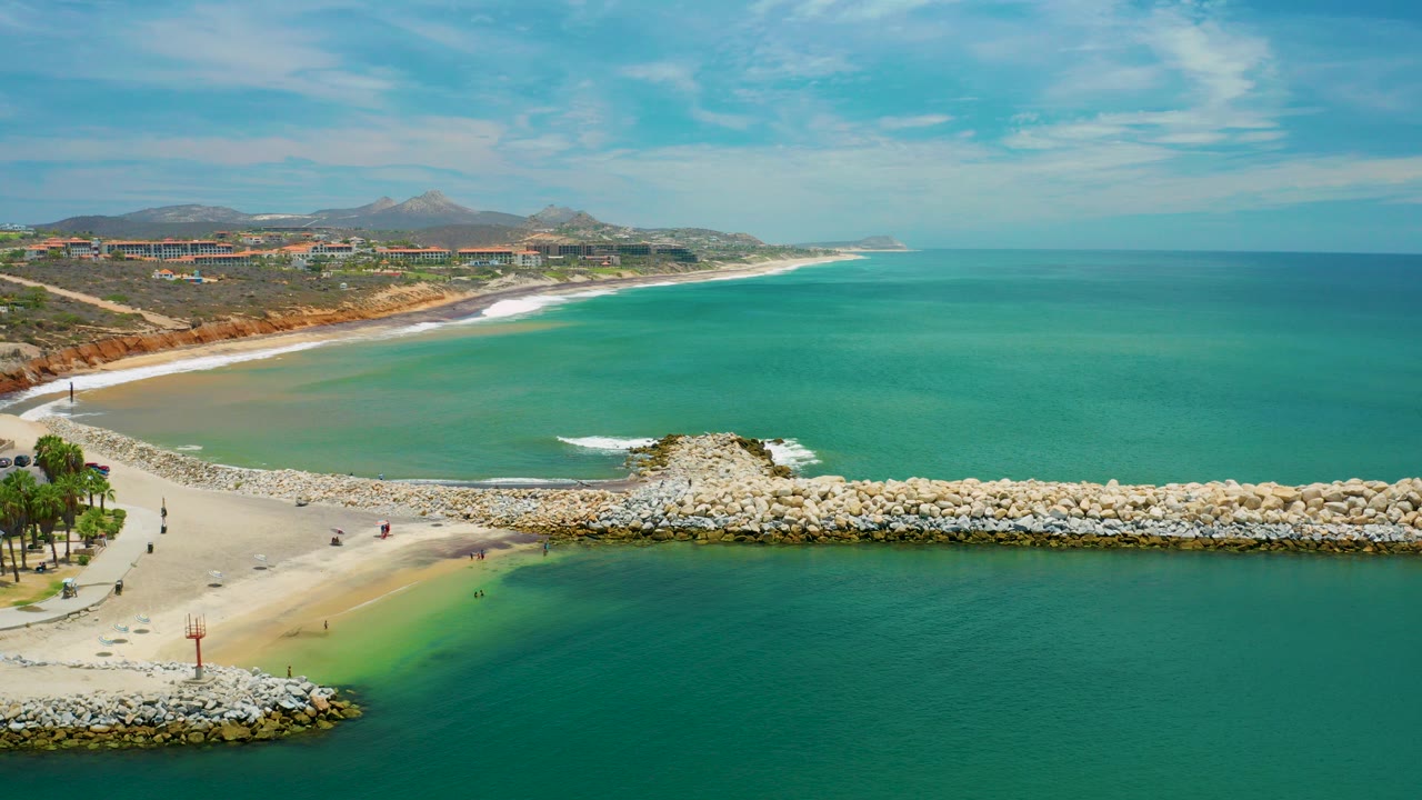 Aerial view in San Jose del Cabo, Mexico showing turquoise water around a pale sand bar, a rocky breakwater across the frame, distant surf, hills, and no discernible people.