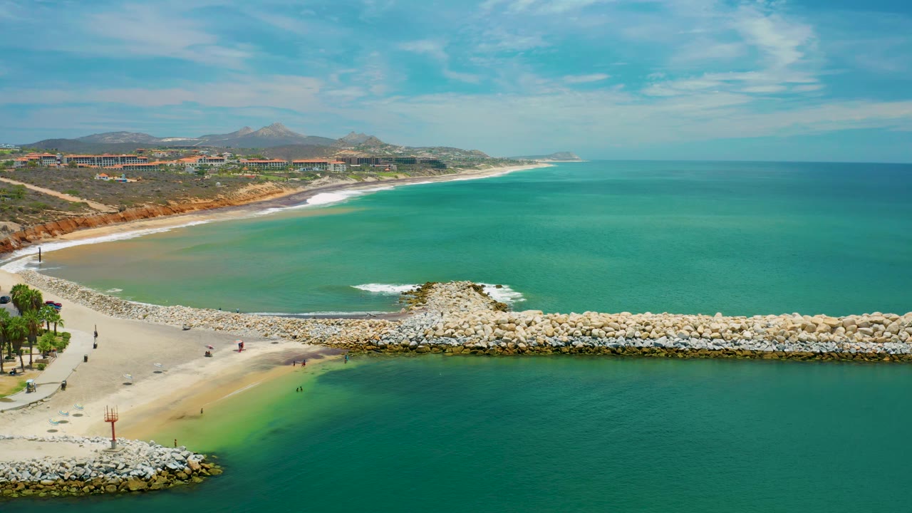 Aerial frame over San Jose del Cabo with calm green-blue lagoon water in the foreground, a pale sand bar at left, a rock barrier through the middle, and no discernible people.