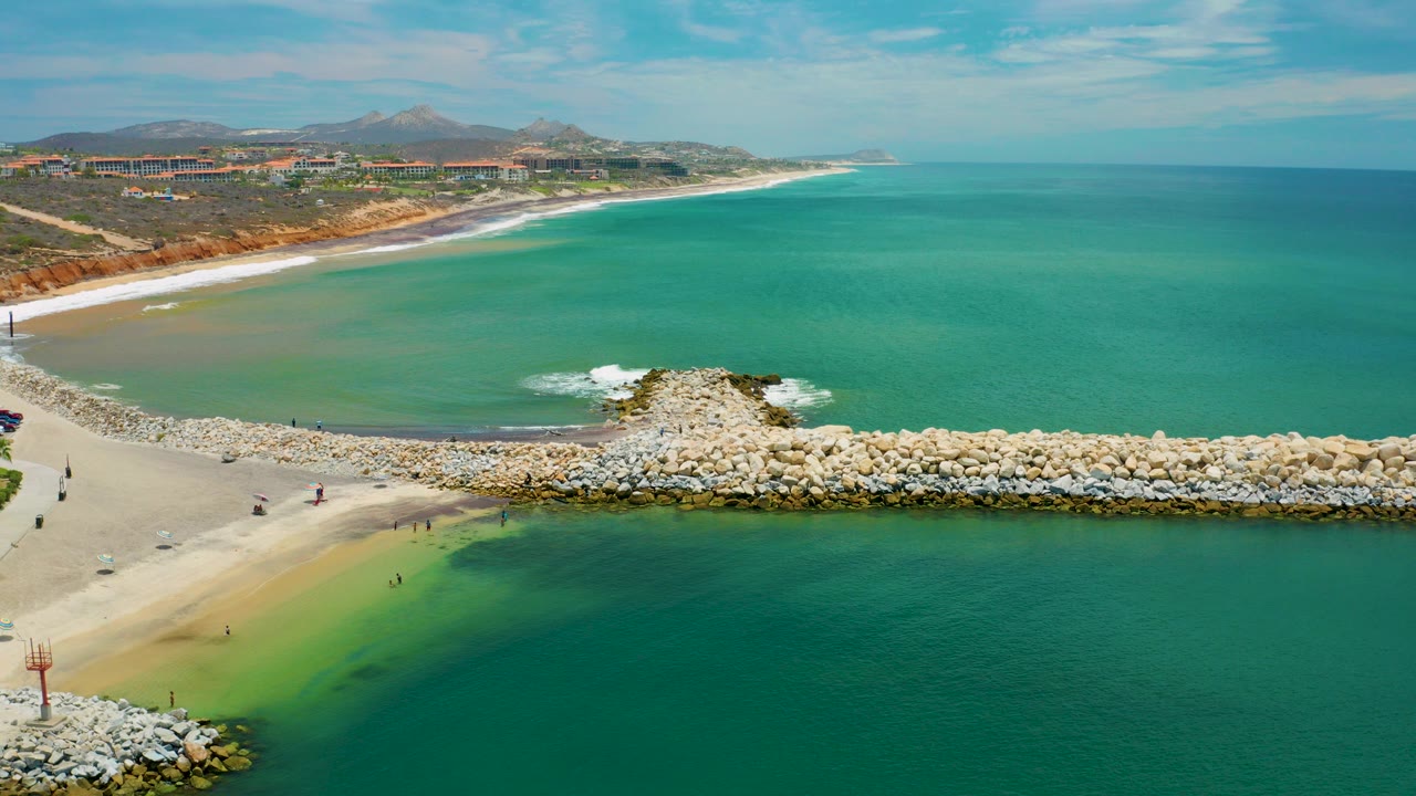 Aerial frame over San Jose Del Cabo water with a rock breakwater crossing the lower half, sandy beach at left, distant hills and buildings, and small visible people on the sand.