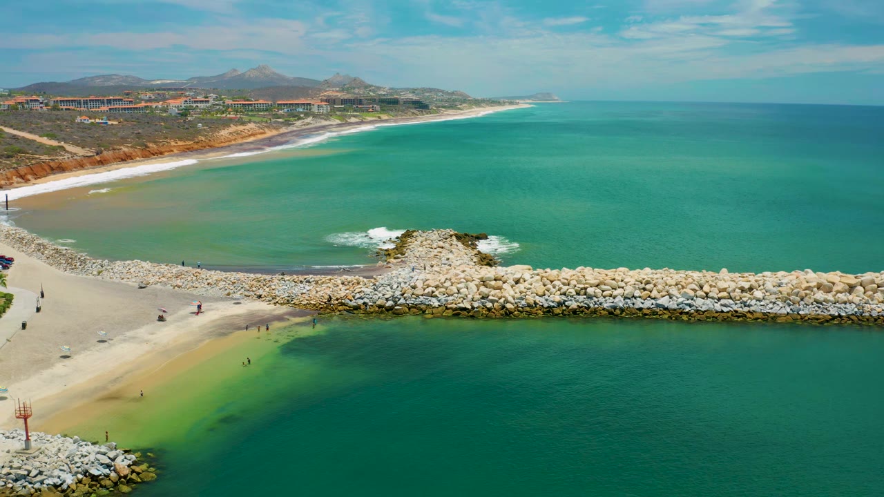 San Jose Del Cabo aerial view with green-blue water in the foreground, pale rocks forming a jetty across the beach, white surf outside the breakwater, and tiny people near the shoreline.