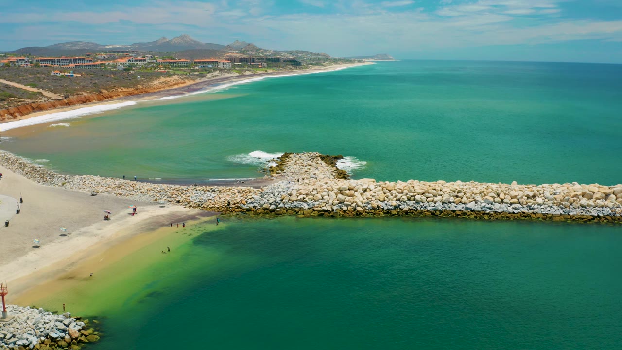 Aerial view in San Jose Del Cabo showing water wrapping around the rock breakwater, a narrow sandy beach at left, surf beyond the rocks, and small figures visible near the waterline.