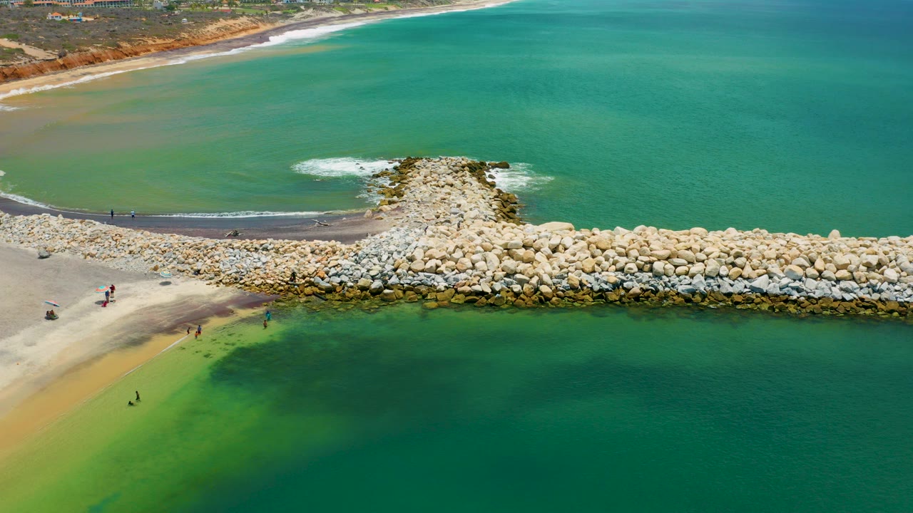 Aerial frame from San Jose Del Cabo, Mexico showing a pale rock jetty extending from sand into green-blue water, with small distant people near the shore.