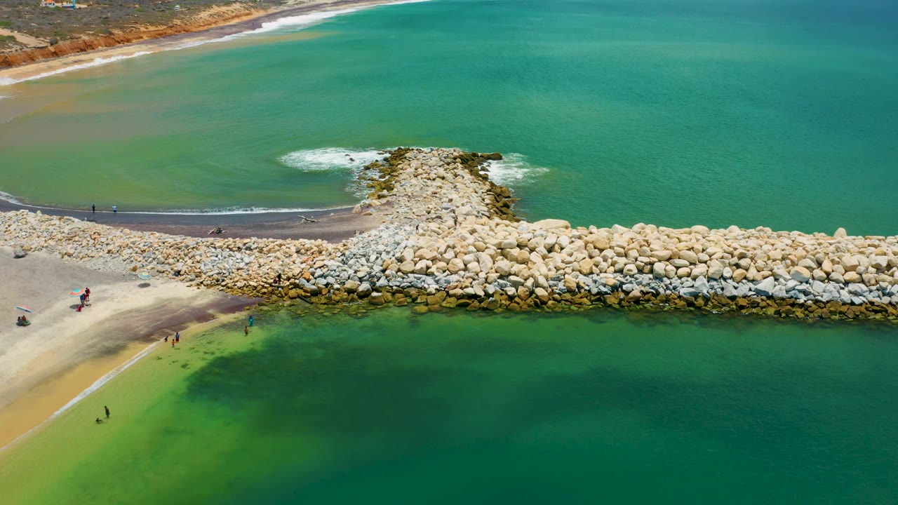 San Jose Del Cabo aerial frame showing the jetty centered below open turquoise water, with a narrow dark beach line and tiny beachgoers along the shore.