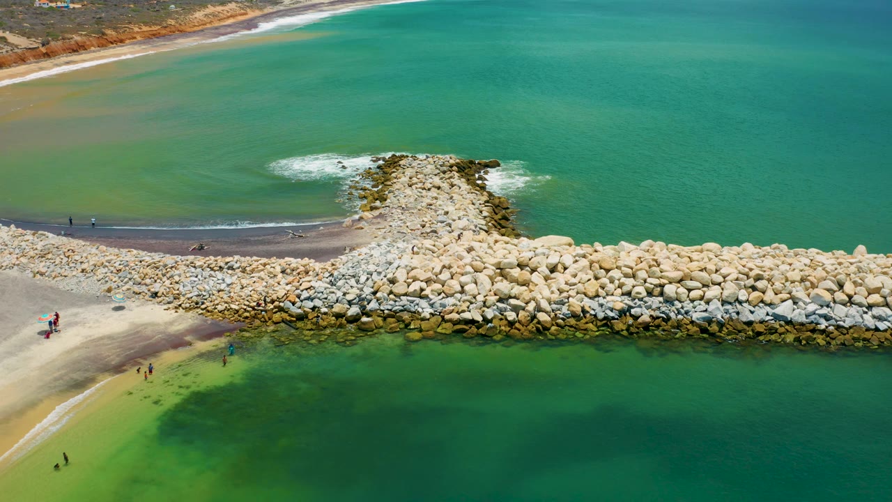 Aerial shoreline with rock jetty, dark sand, and green water below.