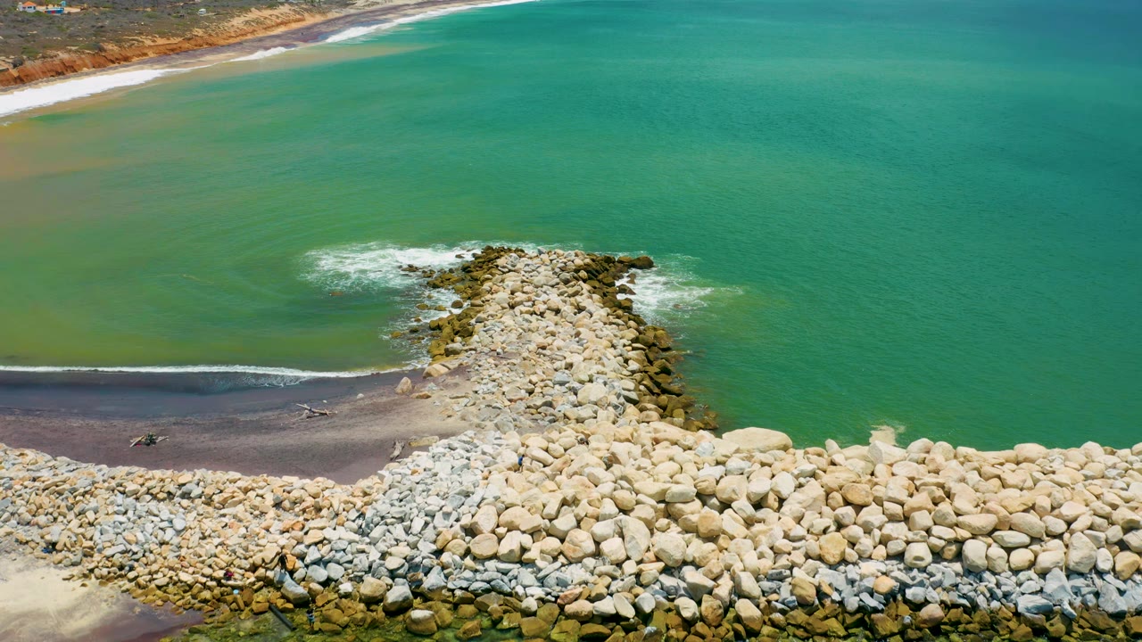 Aerial San Jose Del Cabo frame with the rock jetty centered, pale boulders in the lower foreground, mild white surf on both sides, green water beyond, and no visible people.