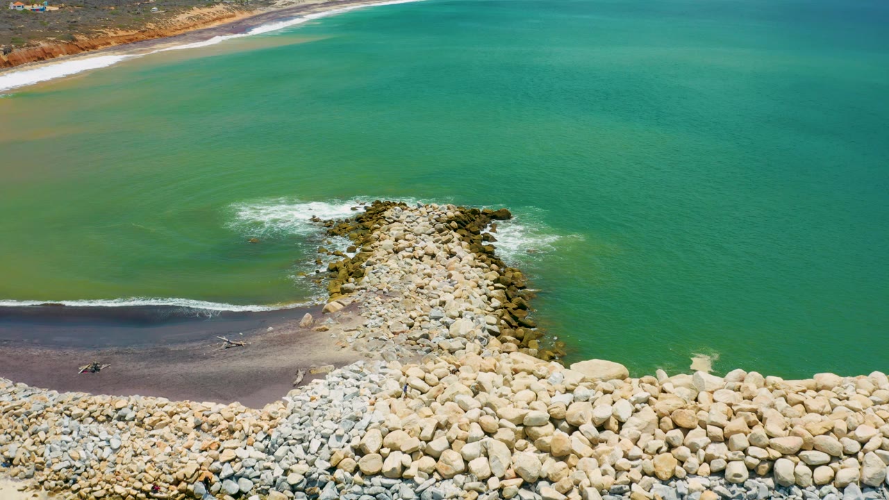Aerial view in San Jose Del Cabo, Mexico with the rock breakwater pushed slightly right of center, turquoise-green water across the frame, a thin beach line near the top, and no visible people.
