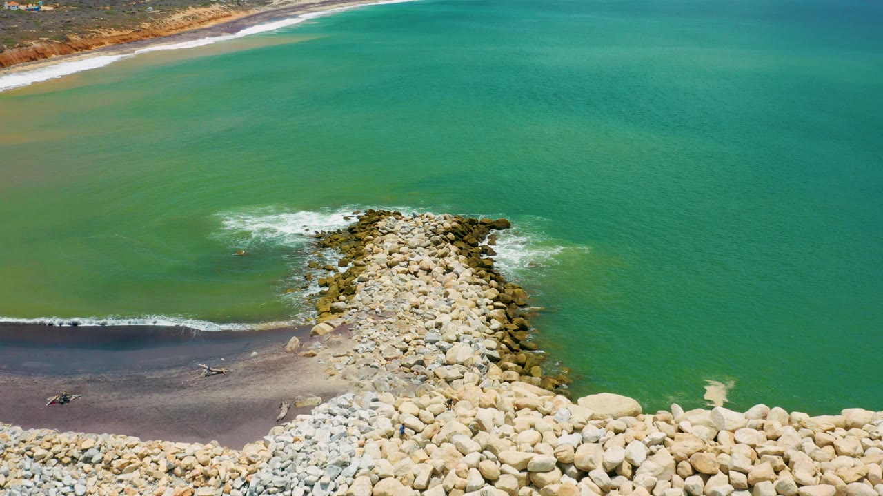 San Jose Del Cabo aerial frame showing the jetty angled into the sea, dark wet sand at lower left, small white waves curling along the shore, green water, and no visible people.