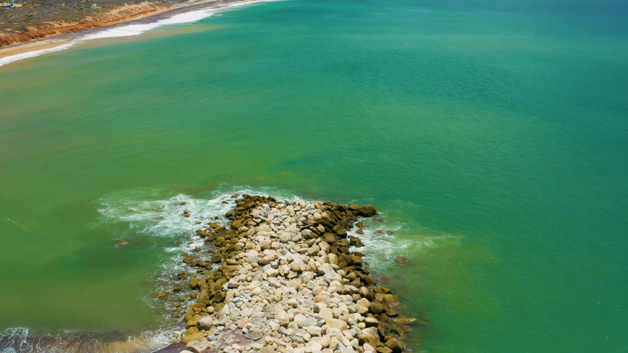 Aerial view in San Jose Del Cabo with the boulder formation near the lower center, a thin beach line at the top left, broader turquoise water to the right, and no visible people.