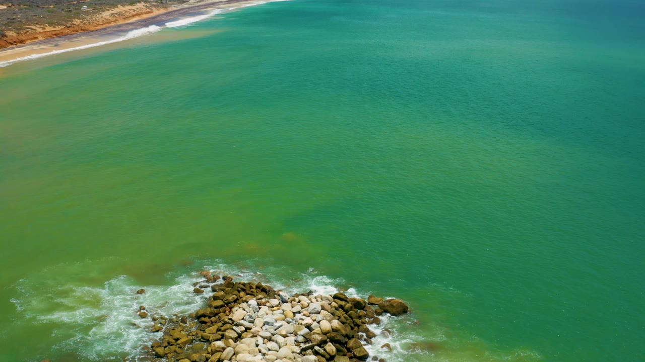 Aerial frame over San Jose del Cabo with blue-green water filling most of the view, white foam around the rocks below, shoreline at the top, and no visible people.