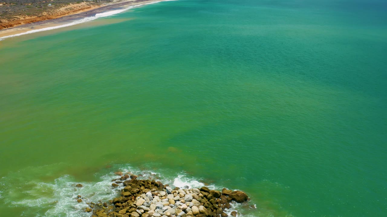 Aerial frame over San Jose del Cabo where the rocky breakwater sits lower in the shot, surf curls around it, the beach stays along the upper edge, and no visible people appear.