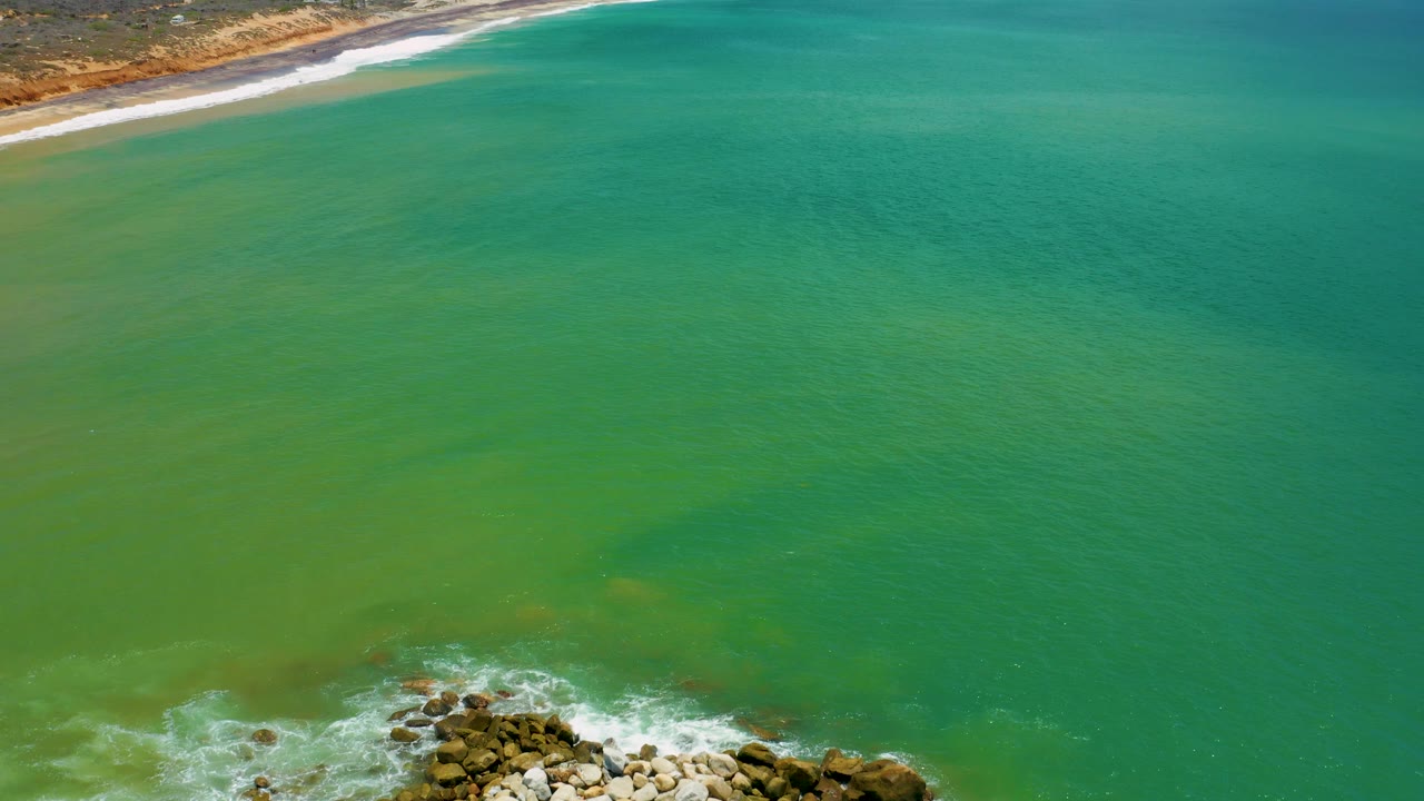 Aerial frame over San Jose del Cabo with broad green water across the center, the rocky breakwater partly cropped at the lower edge, beach at the top, and no visible people.