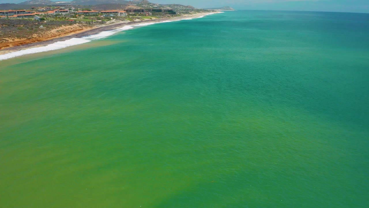 Aerial view over blue-green water off San Jose Del Cabo, Mexico, with a sandy surf line, distant buildings, low hills, and no visible people.