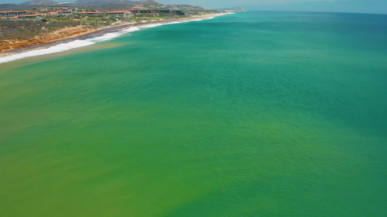 Wide aerial frame of turquoise and green sea near San Jose Del Cabo, with the coastline angled across the upper left and no visible people.