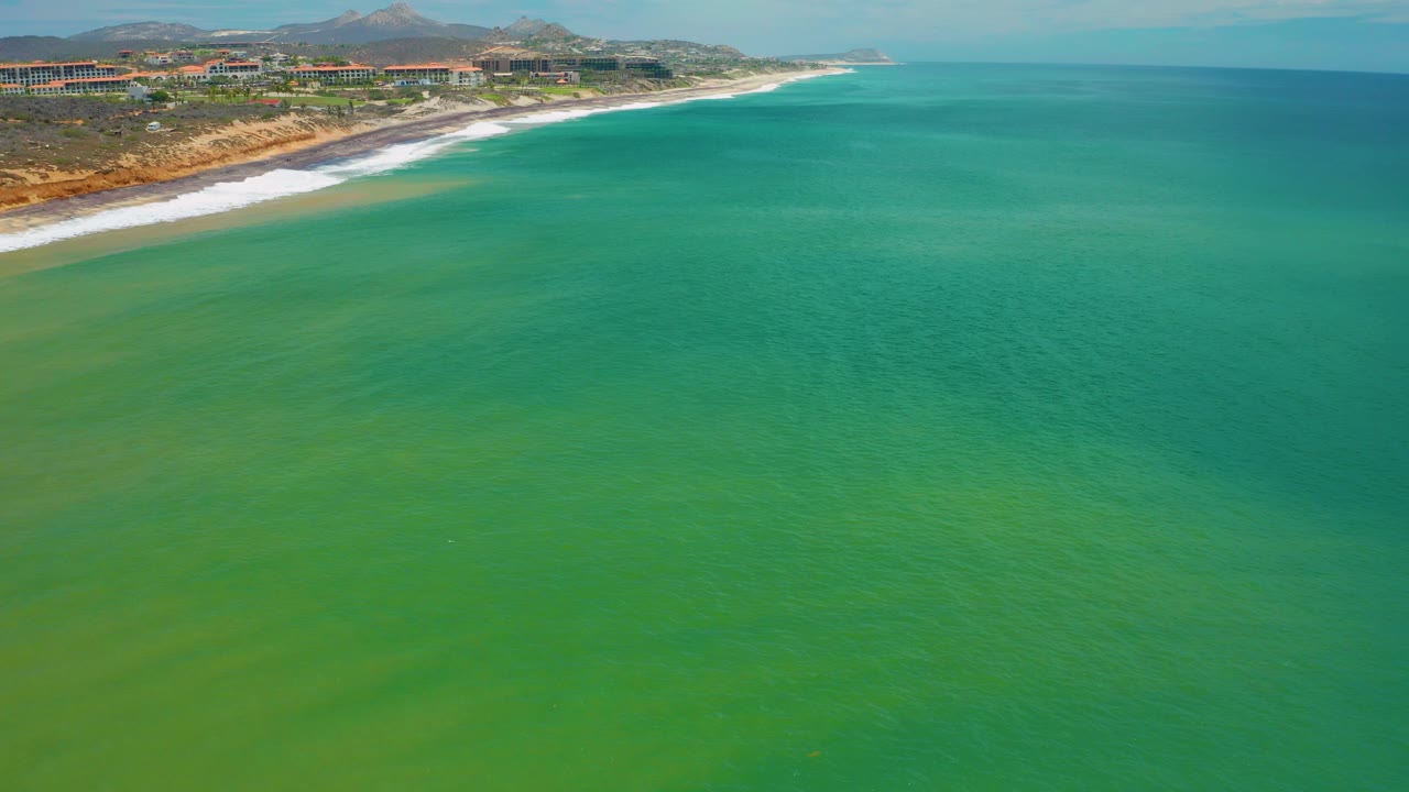 Elevated view of blue-green ocean at San Jose Del Cabo, Mexico, with a white surf band, shoreline buildings, hills, and no visible people.