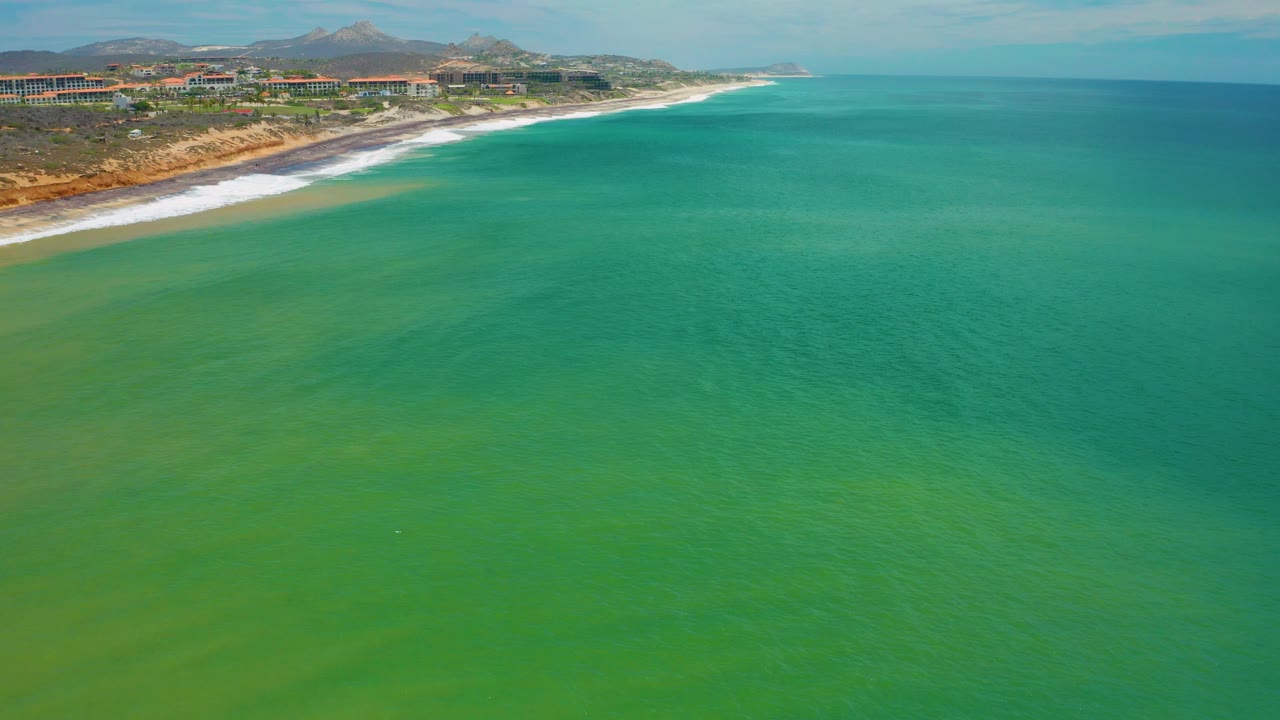 Aerial frame over green-blue water in San Jose Del Cabo, with the sandy coast and hillside background shifted along the upper left and no visible people.
