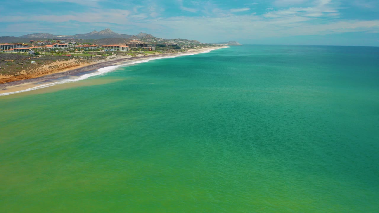 Aerial frame over San Jose Del Cabo, Mexico showing emerald water, a left-edge surf line, beach, coastal buildings, brown hills, blue sky, and no visible people.