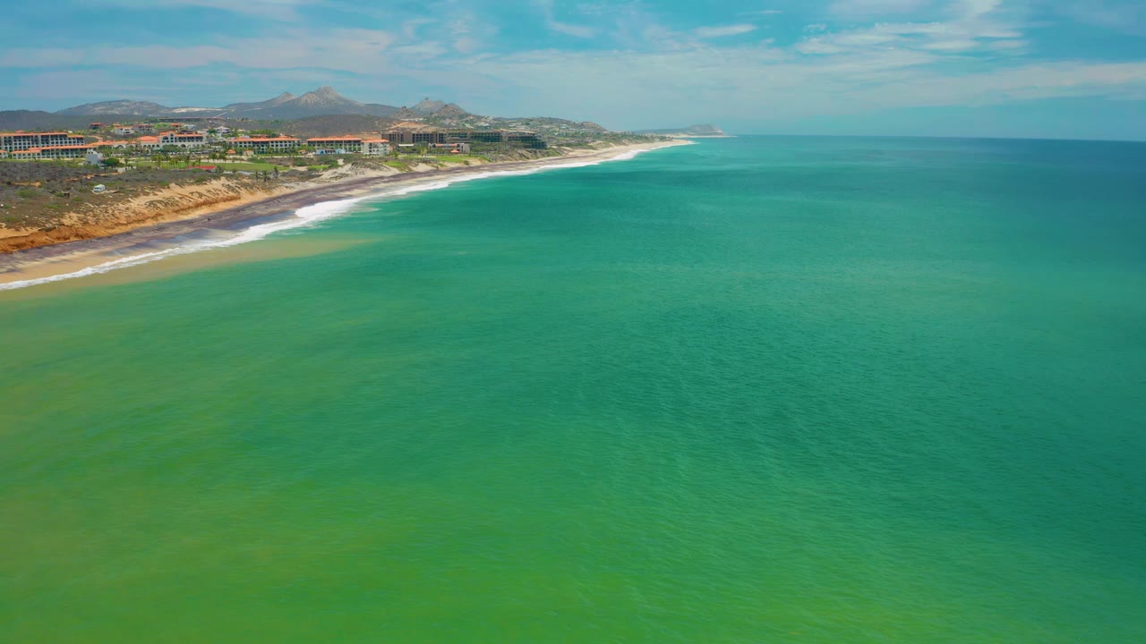 Aerial view in San Jose Del Cabo, Mexico with open green ocean across the foreground, a narrow beach and white surf at left, low buildings near shore, hills behind them, and no visible people.