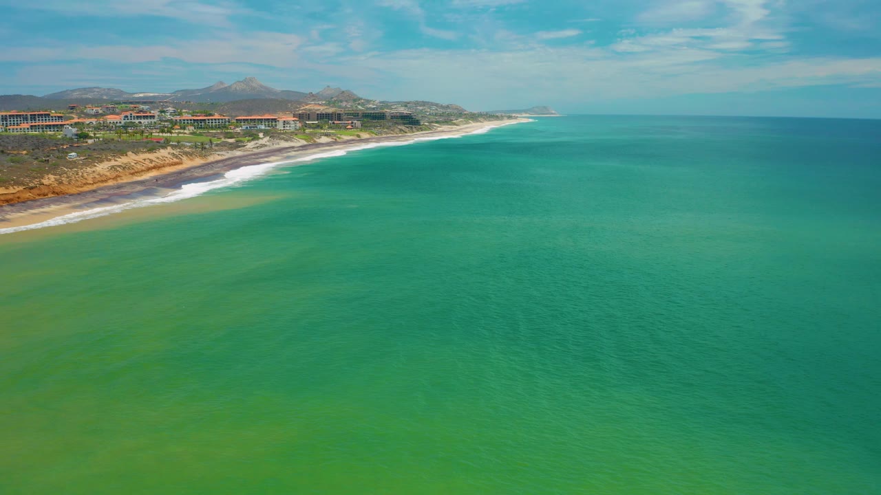San Jose Del Cabo, Mexico coastline seen from above, with green water dominating the frame, a curving band of surf and sand at left, buildings on the bluff, distant hills, and no visible people.