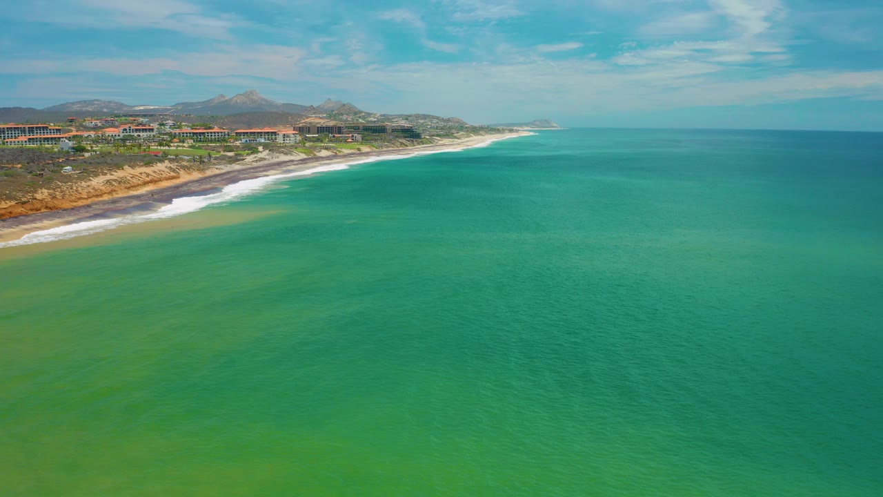 Aerial San Jose Del Cabo, Mexico frame showing green sea in the foreground, white surf tracing the beach at left, hillside coastline in the distance, coastal buildings, and no visible people.