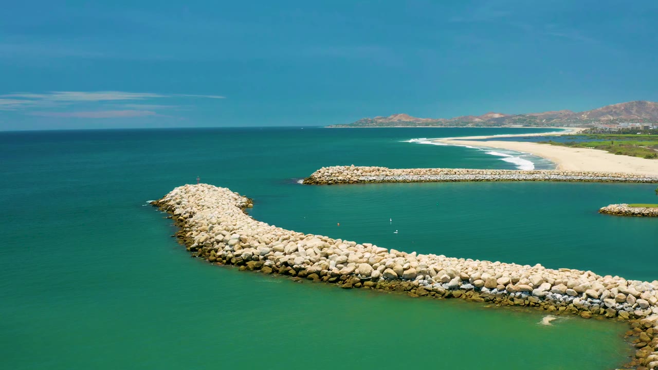 Second frame from San Jose del Cabo, Mexico, with the rock jetty arcing across the foreground, calm ocean filling the center, a white surf line on the beach, and no visible people.