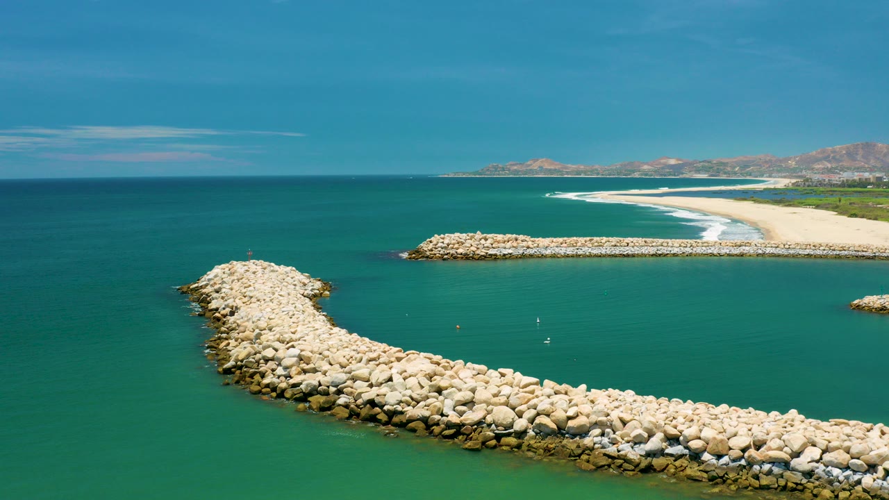 Opening aerial view in San Jose Del Cabo, Mexico, with a curved boulder breakwater in the foreground, turquoise water around it, beach surf in the distance, and no visible people.